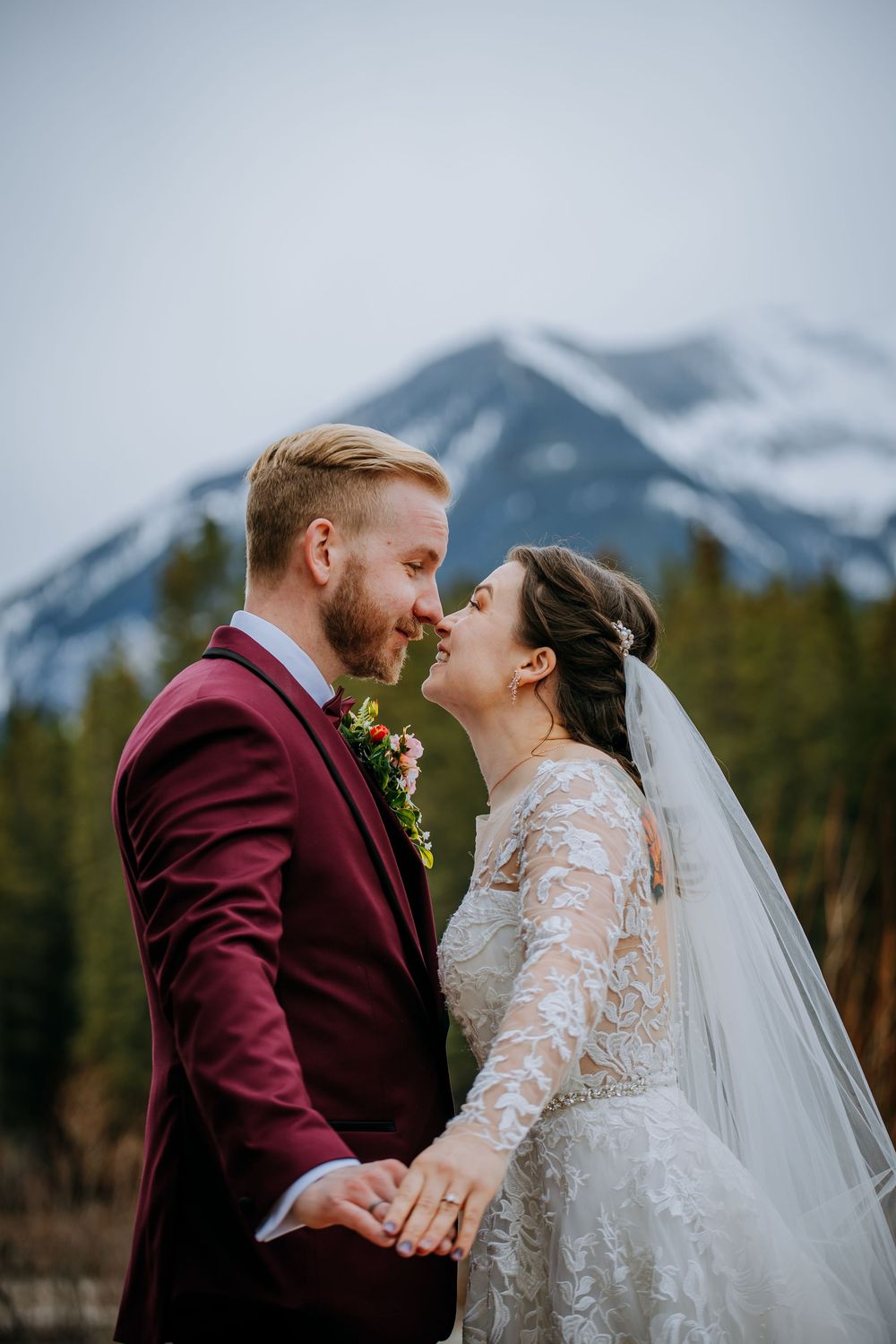 Bride and groom nearly kissing with snowy mountain backdrop, groom in burgundy suit, bride in lace wedding dress with veil.