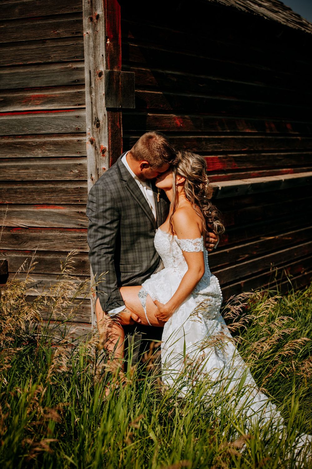 Romantic wedding couple embraces by rustic wooden barn wall in tall summer grass at sunset.
