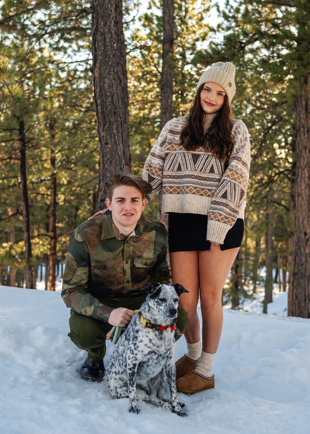 Couple and their spotted dog pose together in snowy winter forest setting.