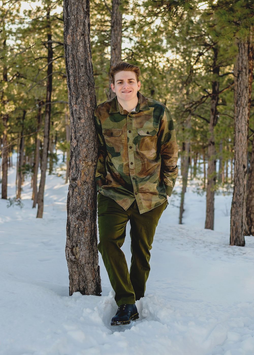 Person in camouflage jacket leans against pine tree in snowy forest setting.
