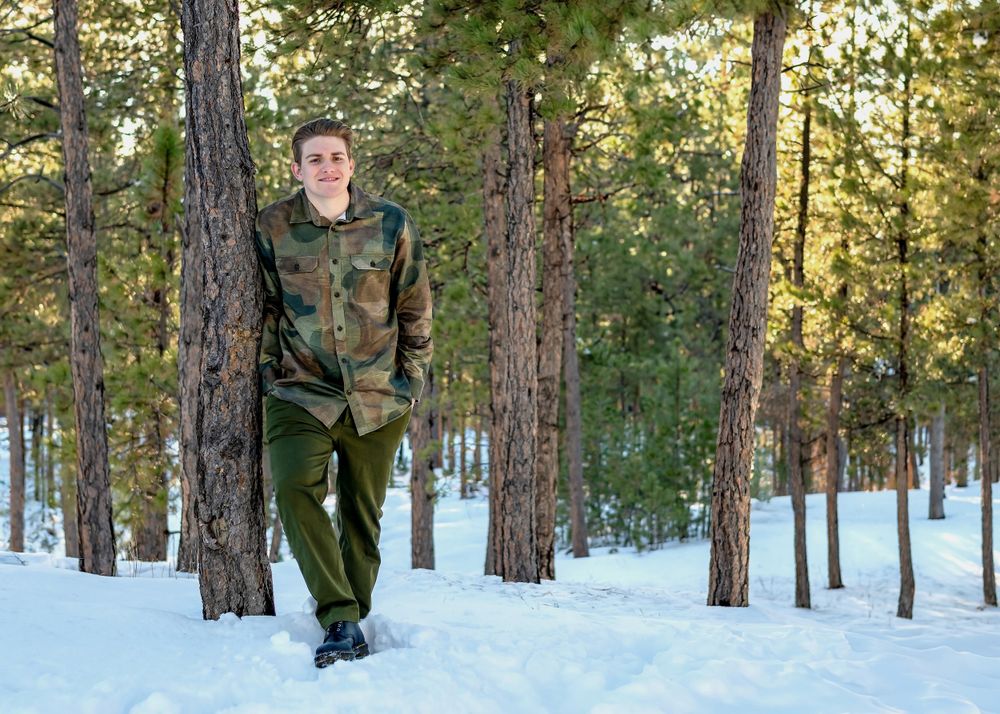 Person in camouflage clothing standing among snow-covered pine trees.