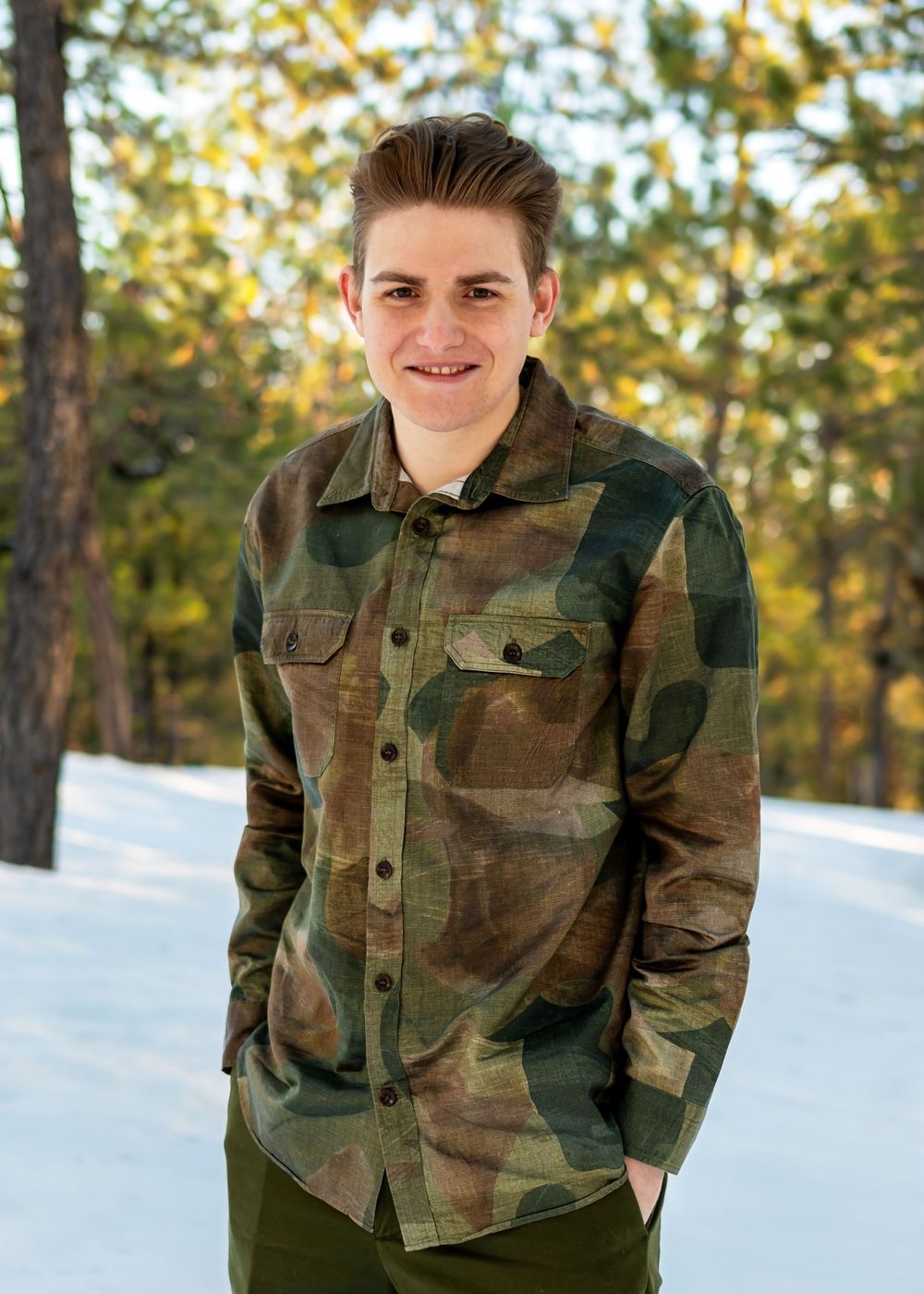 Young man in camouflage jacket smiling outdoors in snowy forest setting.