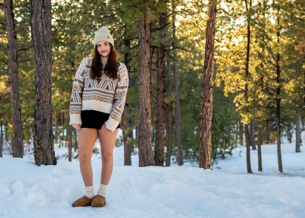Woman in a patterned sweater and beanie poses in a snowy forest during golden hour sunset.