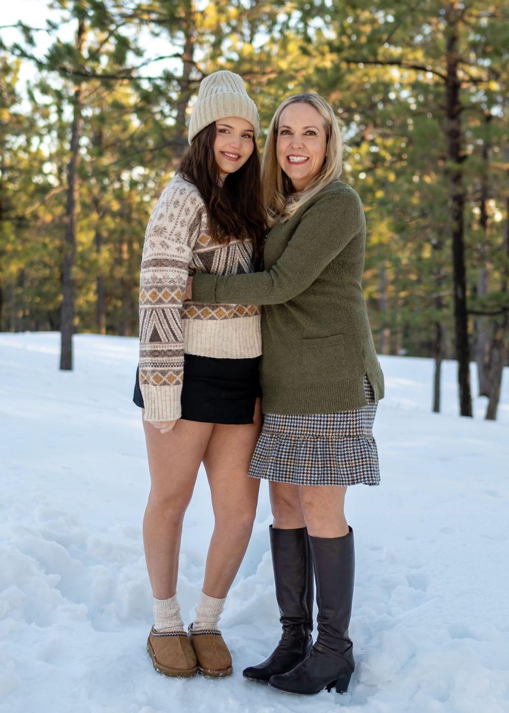 Two people embrace while standing in snowy forest wearing winter outfits.
