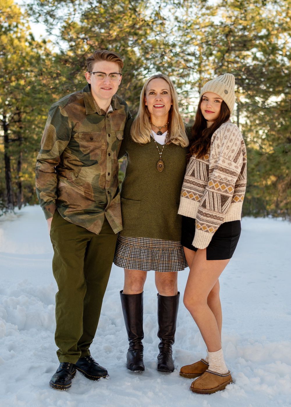 Family in winter attire poses together in a snowy forest setting wearing sweaters and boots.