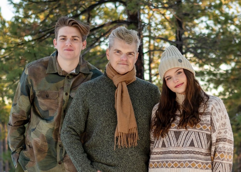Three people in cozy winter clothing pose together in a snowy forest with pine trees in the background.