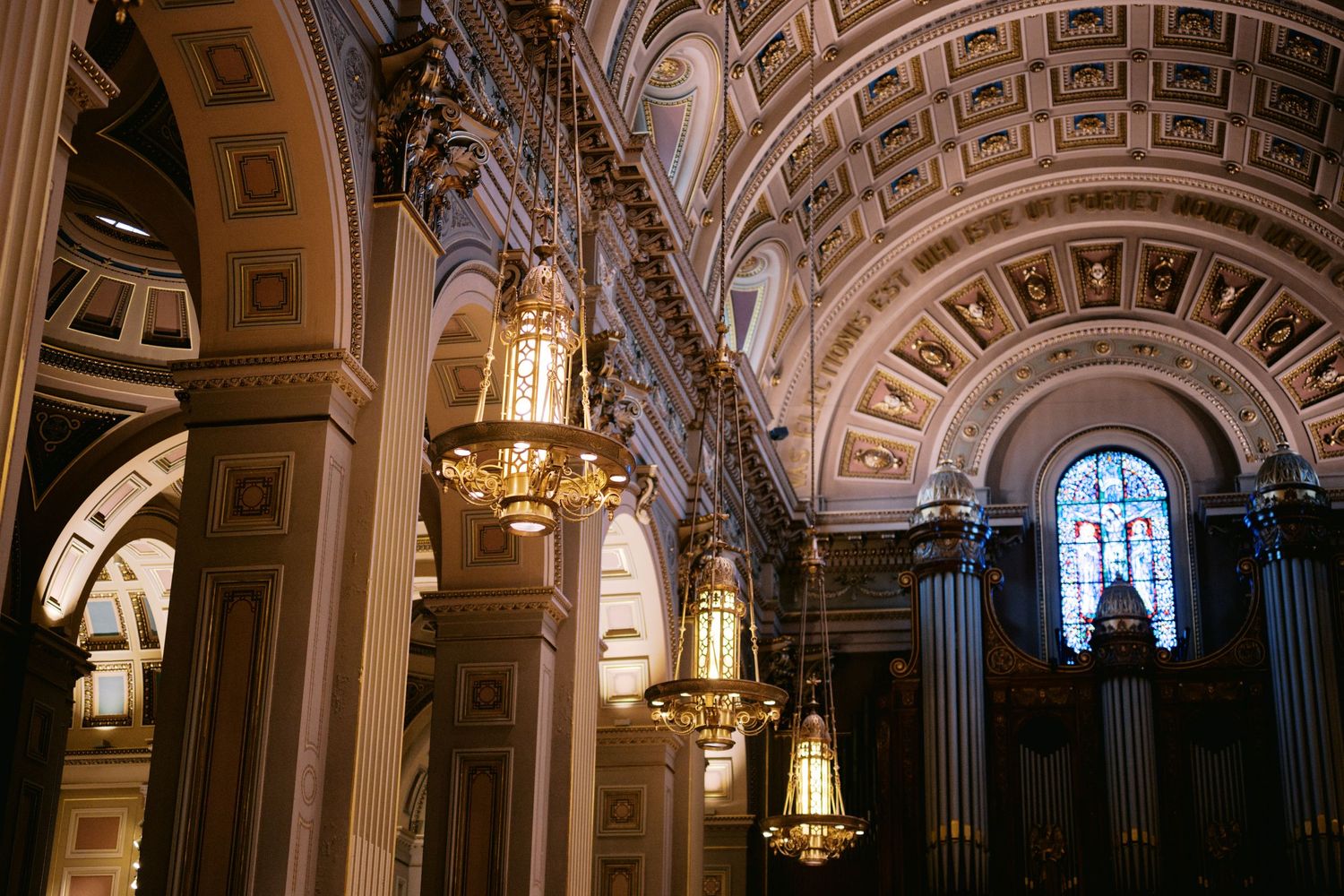 Ornate cathedral interior with hanging brass lanterns and arched baroque ceiling details illuminated by warm lighting.