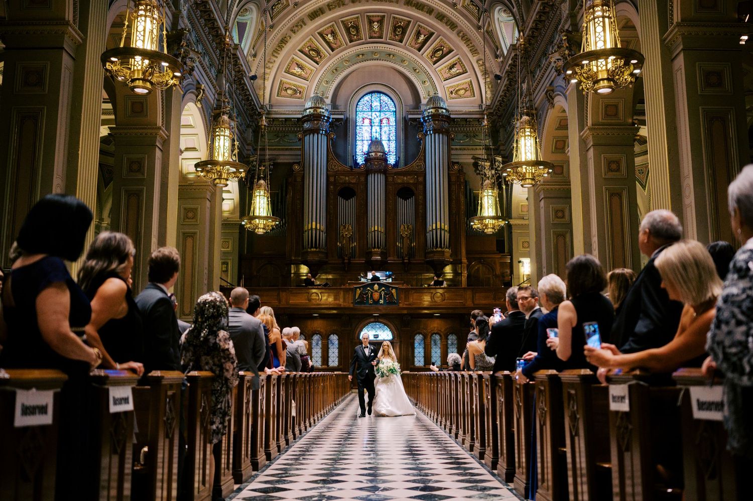 Wedding ceremony inside a grand cathedral with ornate architecture, hanging chandeliers and guests seated in wooden pews.