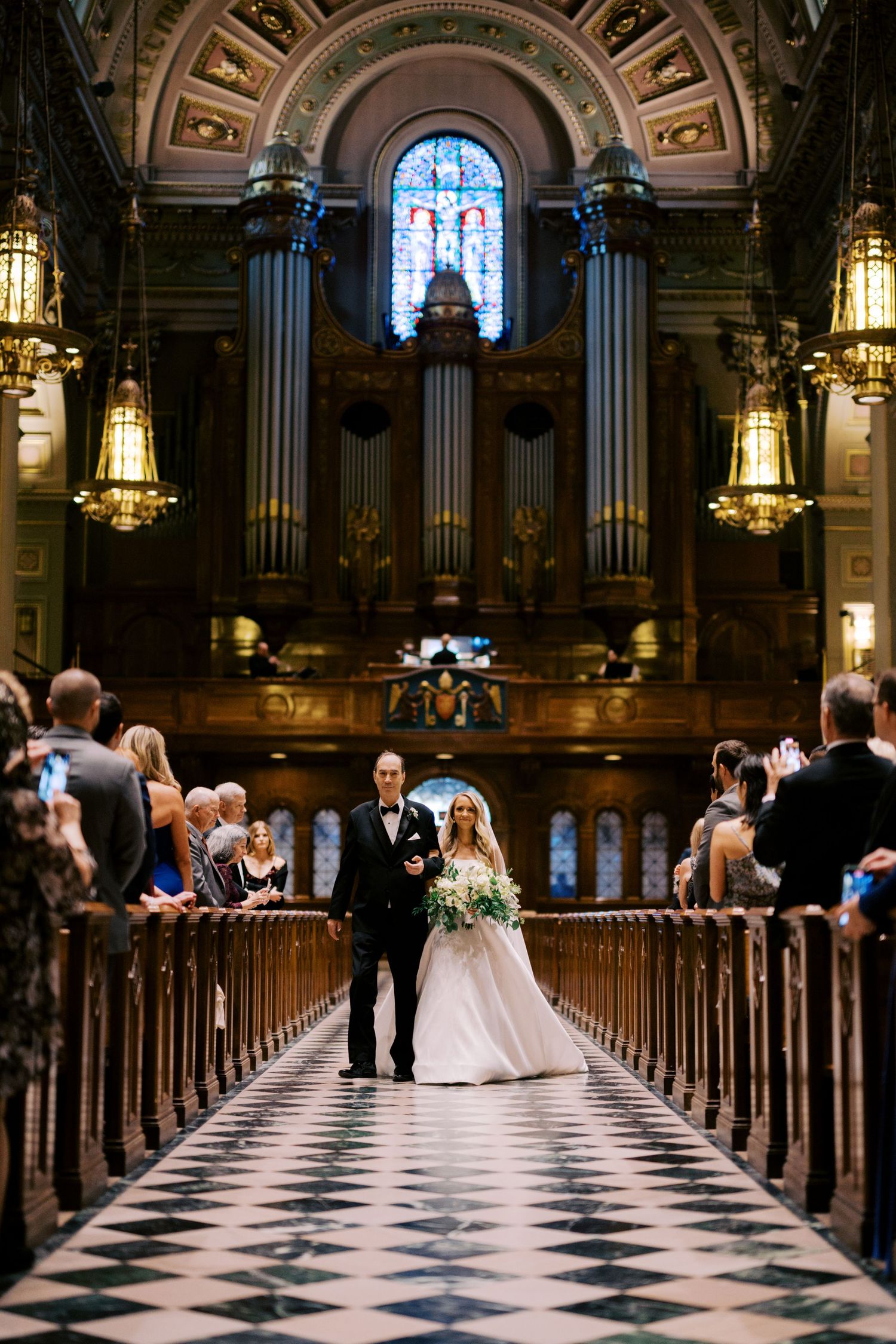 A wedding ceremony in a grand cathedral with ornate pipe organs, hanging lanterns, and a black and white checkered aisle.