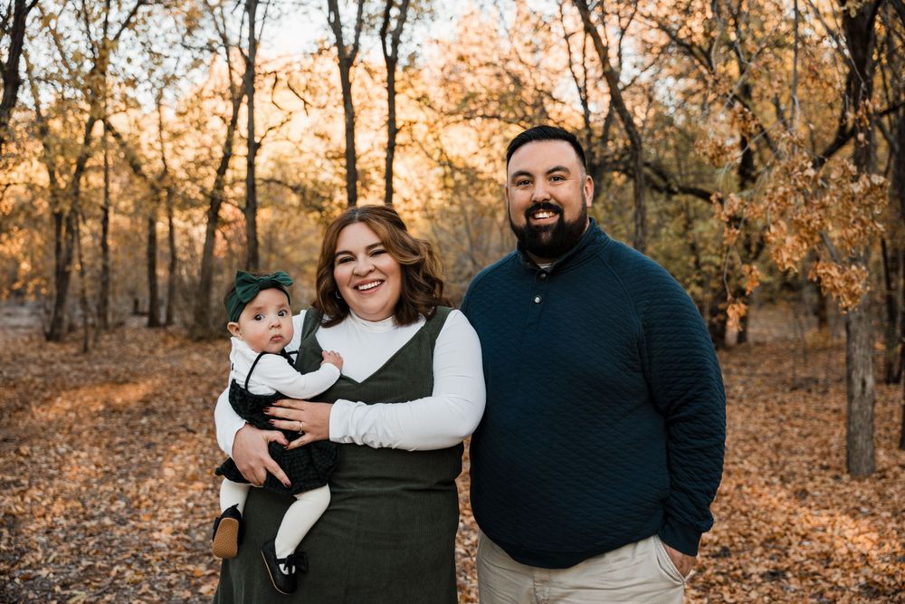 Family of three poses together during fall photo session with orange foliage in background.