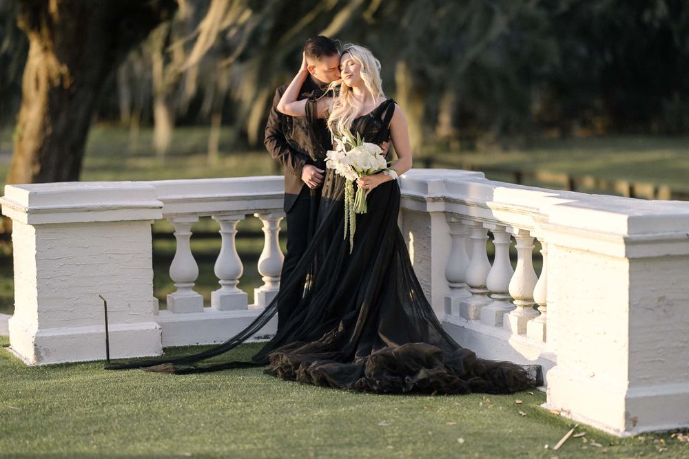 Woman in elegant black gown posing gracefully on white stone bridge balustrade in outdoor garden setting.