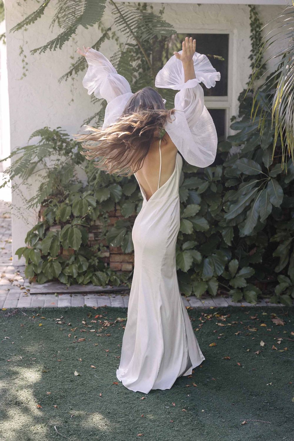 A figure in a flowing white dress dances gracefully in a garden setting with trailing fabric creating dynamic movement.