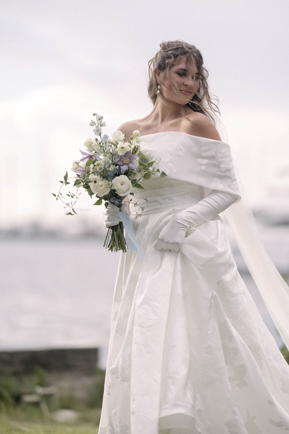 Winter bride in off-shoulder wedding gown holds delicate bouquet of white and green florals against misty backdrop.