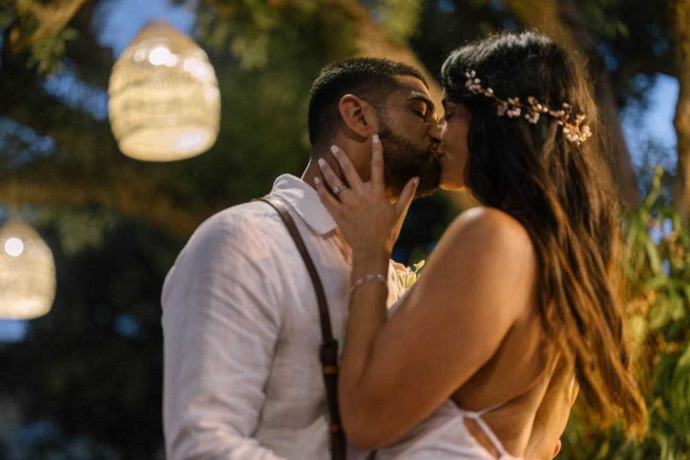 A couple shares a romantic kiss at dusk with glowing lanterns and palm trees in the background.