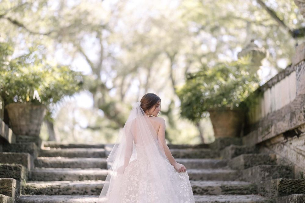 A bride in a flowing lace wedding dress stands on stone steps surrounded by dreamy garden scenery.