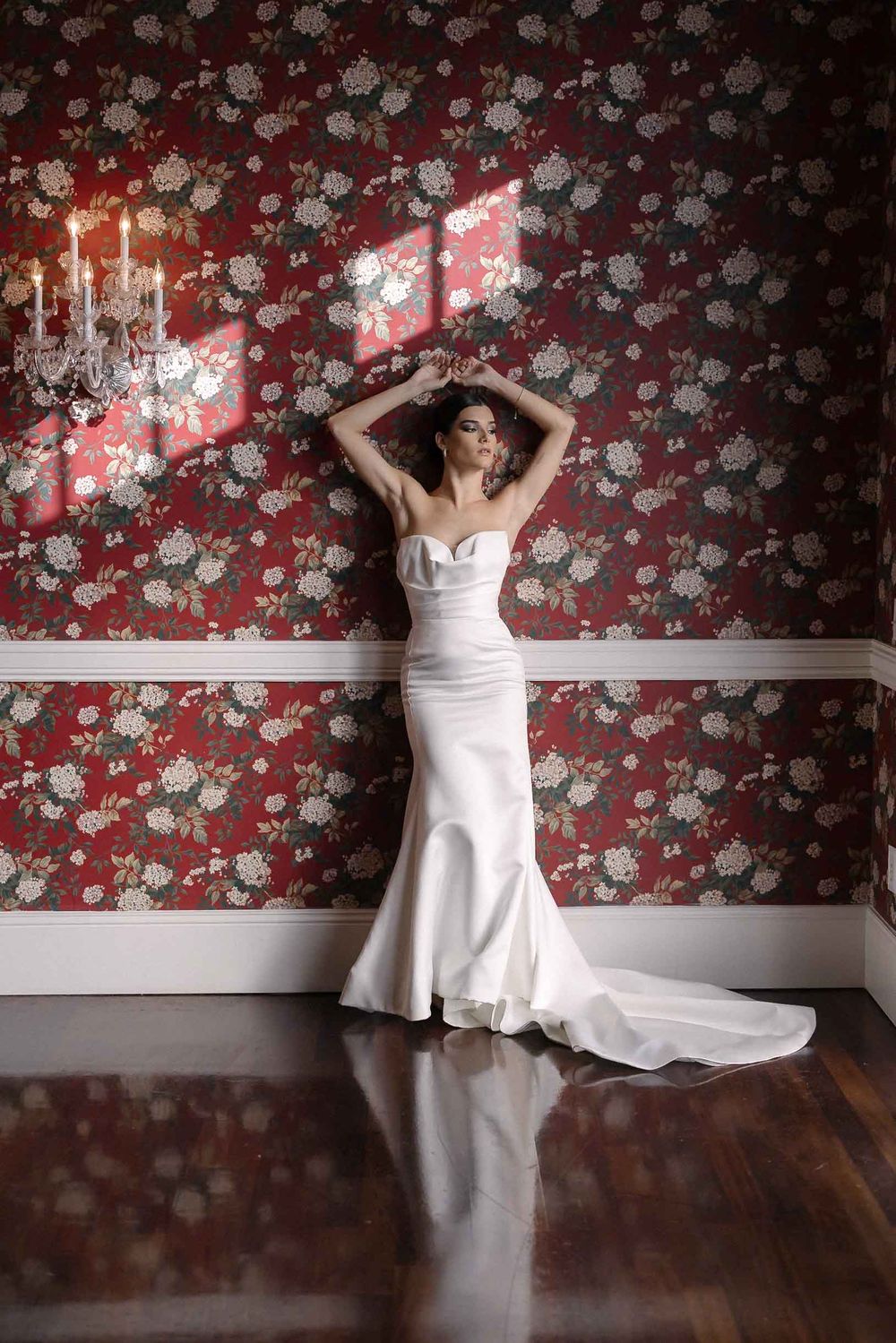 Elegant white mermaid wedding dress against floral wallpaper with candlelit wall sconces in luxurious interior setting.