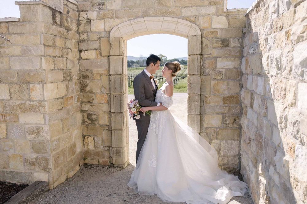 A newlywed couple poses in a stone archway with natural light filtering through during their wedding photoshoot.