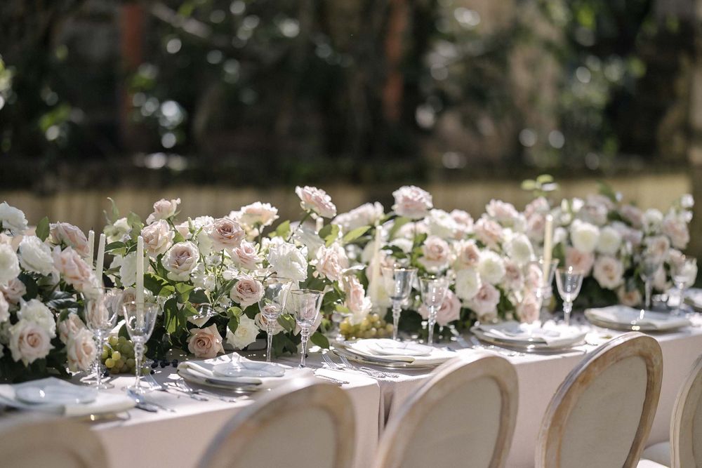 Elegant outdoor wedding reception table set with white chairs and lush floral centerpieces featuring soft pink roses.