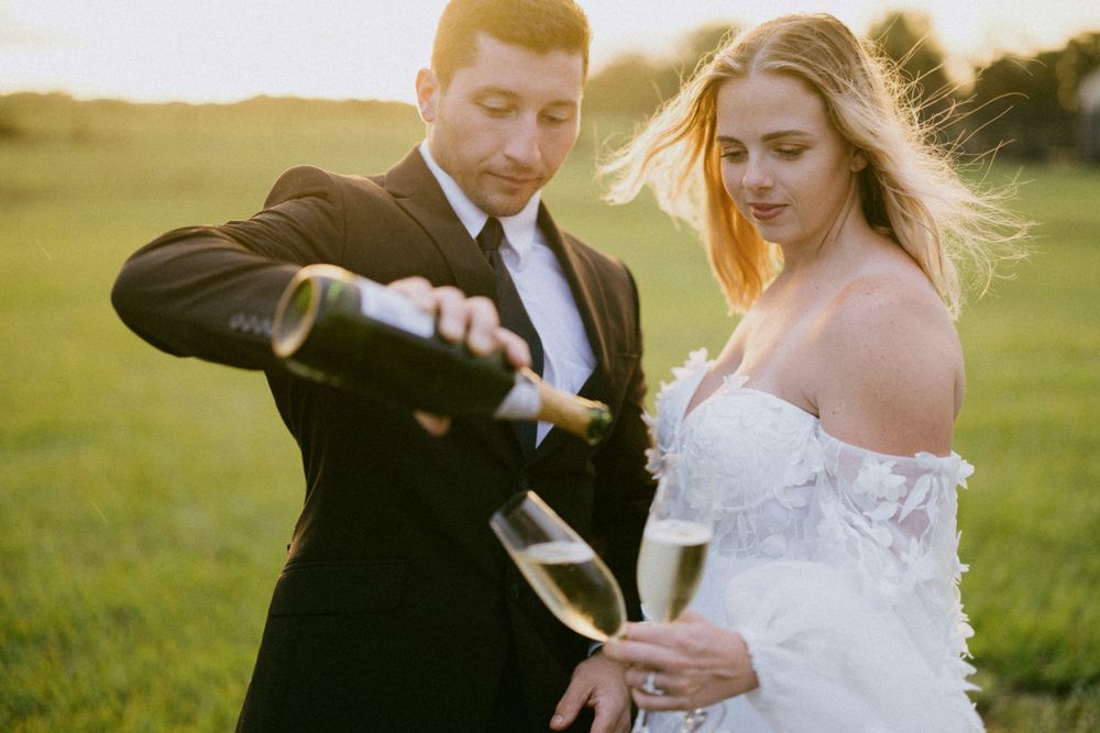 Couple pouring champagne together in formal wedding attire on grassy field.