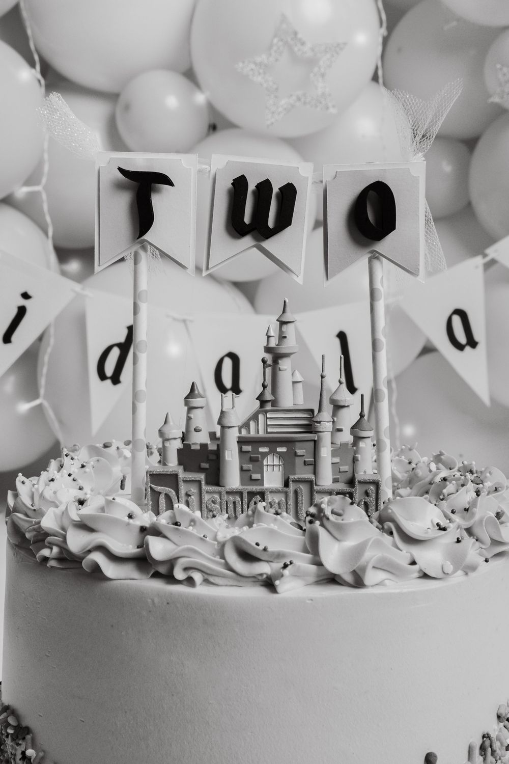 Black and white close-up of castle cake topper with 'Two' banner and white balloons.