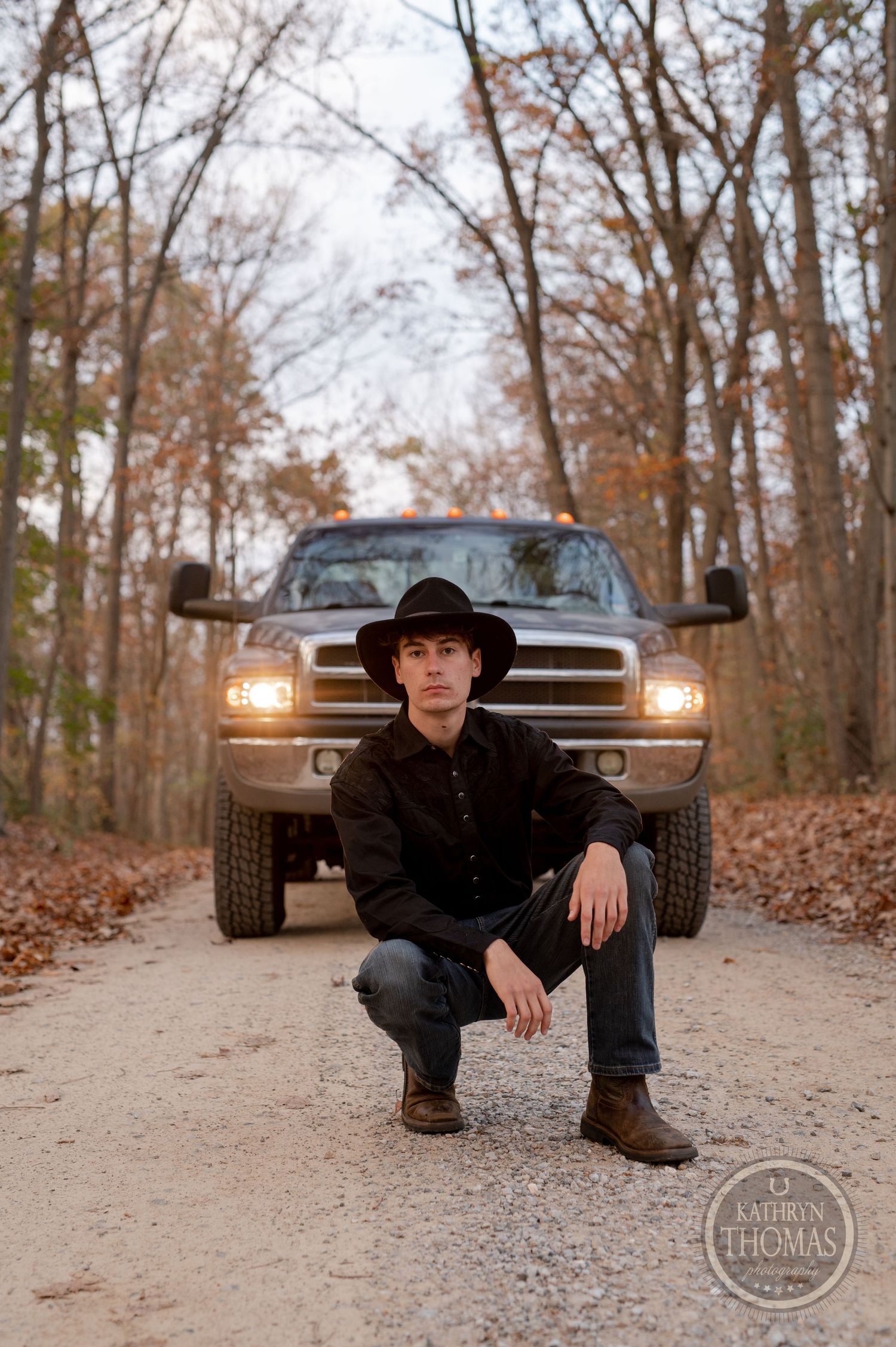 Someone in western wear crouches in front of a pickup truck on a leaf-covered forest path.