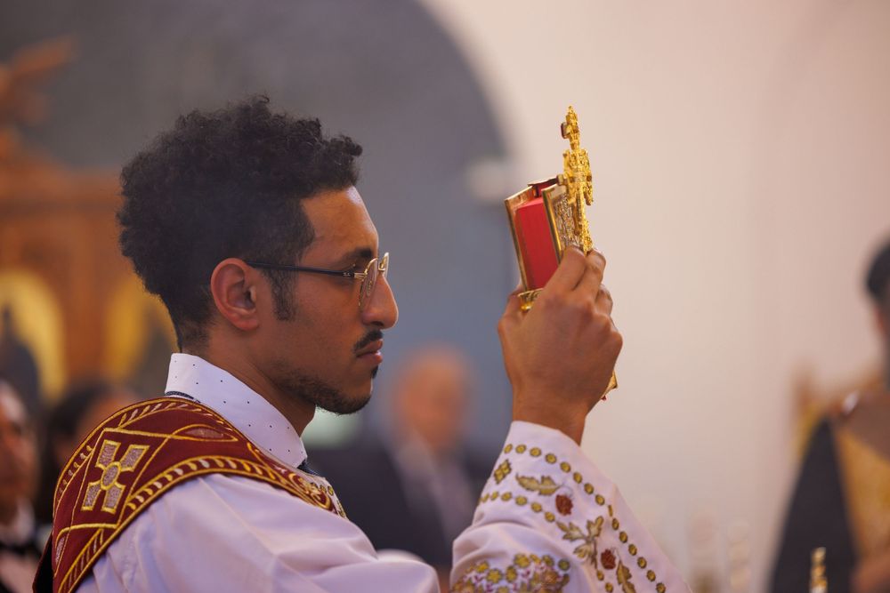 Young deacon holding ornate golden gospel book during Coptic Orthodox Christian religious church service.