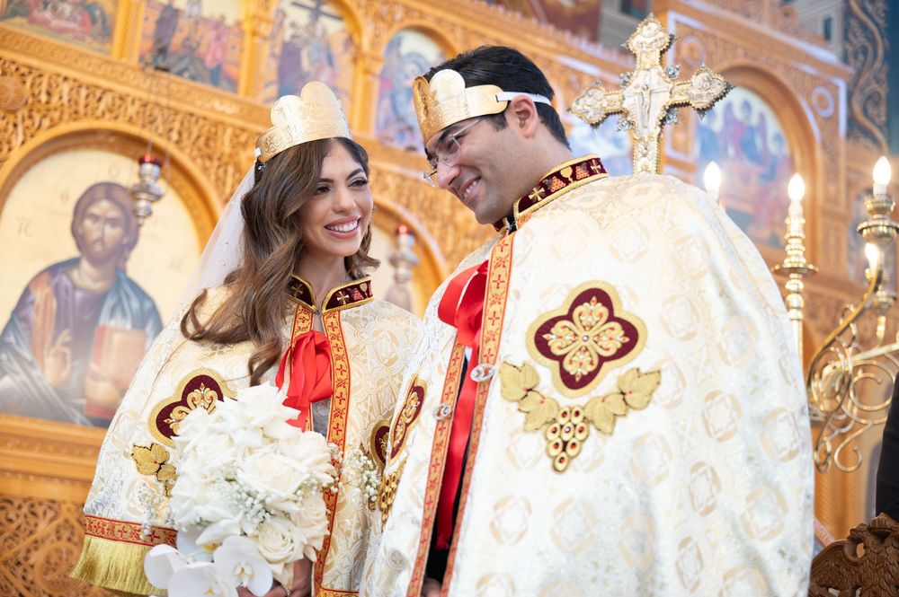 Bride and groom in ceremonial crowns smile during Coptic Orthodox wedding in ornate church.