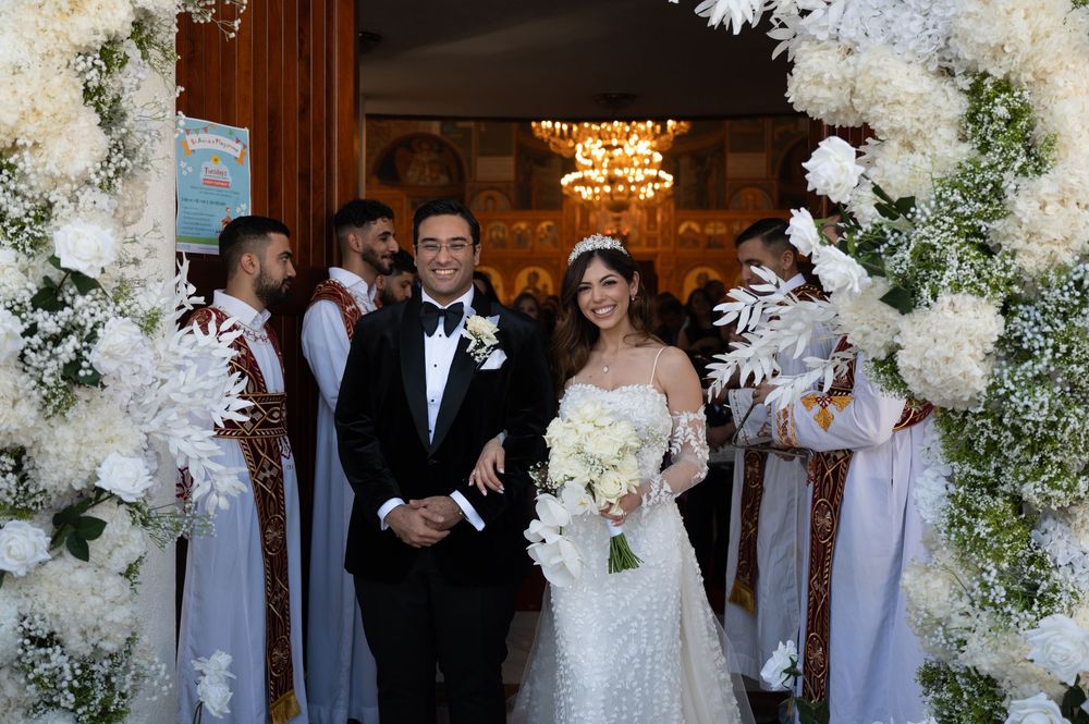 Newlyweds exit church through white floral arch after Coptic Orthodox wedding ceremony.