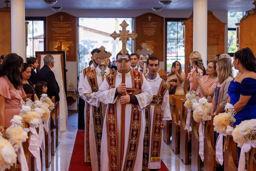 Orthodox priests lead wedding procession down church aisle carrying ceremonial cross and icons.