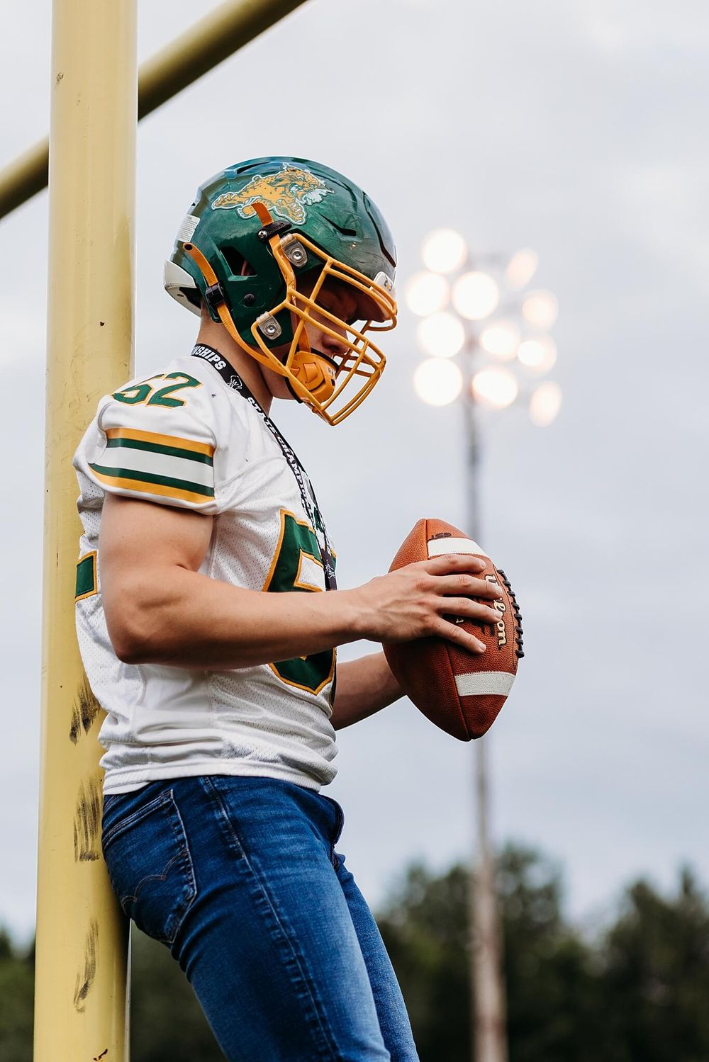 Football player in green and white uniform holds ball next to goalpost at night.