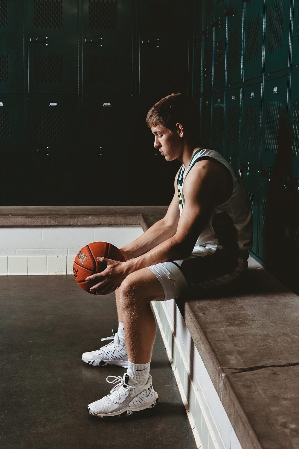 Basketball player sits on bench holding ball in dark locker room with dramatic lighting.