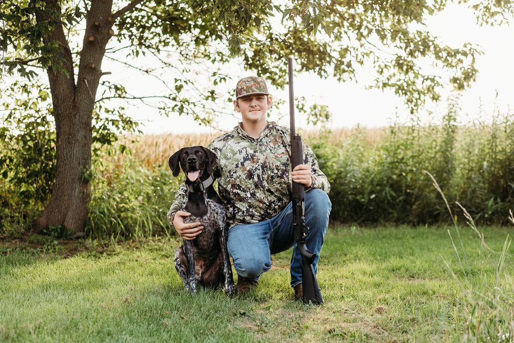 A hunter in camouflage gear poses with a black hunting dog under a tree in a grassy field.