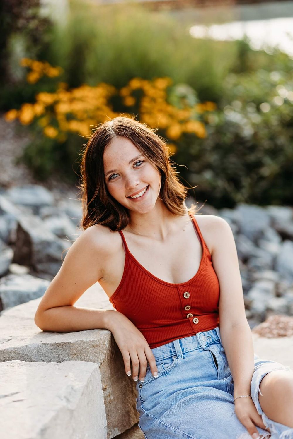 Summer portrait of person in red tank top and denim shorts sitting on stone wall with yellow flowers.