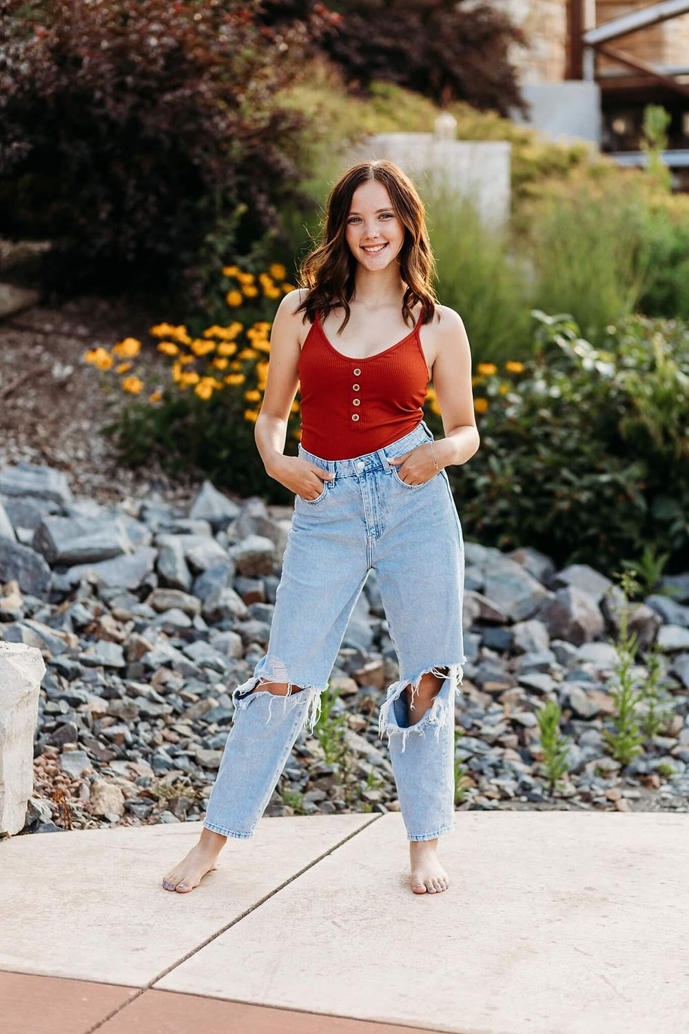 Casual summer outfit with red tank top and distressed jeans against garden background.