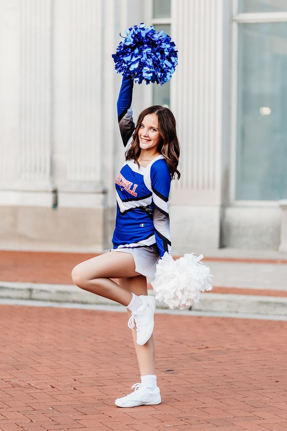 Cheerleader in blue and white uniform holds pom pom while posing on brick surface.