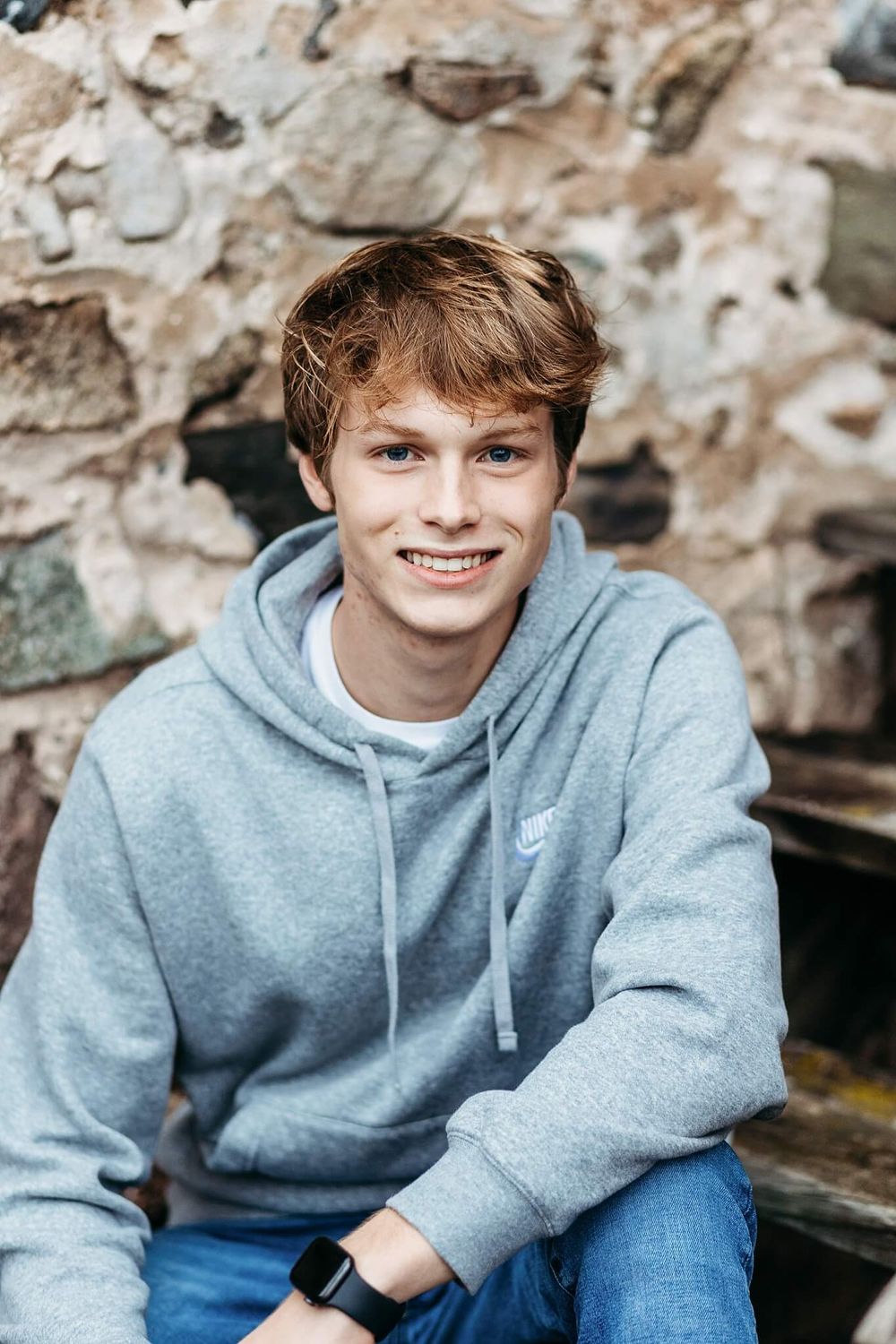 Casual portrait of person in grey Nike hoodie sitting against stone wall.