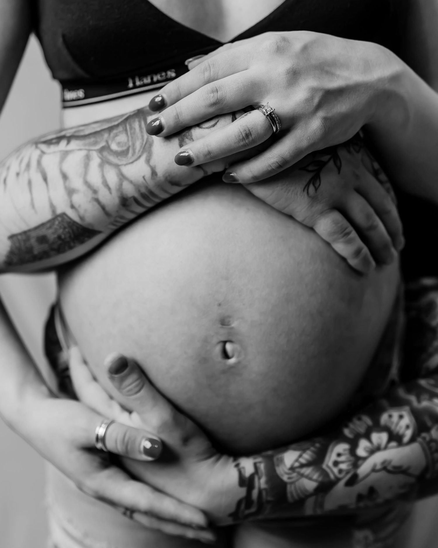 Black and white photo of decorated arms cradling a pregnant belly with rings and tattoos visible.