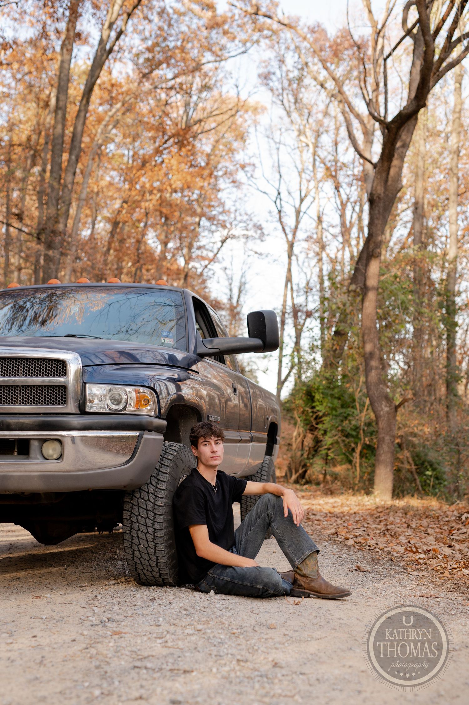 Person sitting against large tire of Dodge pickup truck on leaf-covered forest road.
