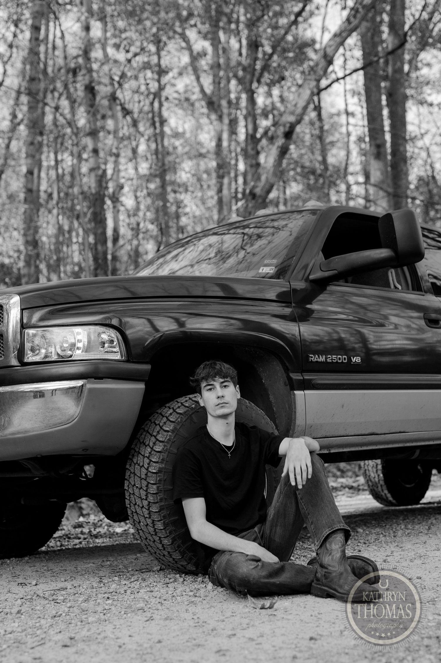 Black Ram pickup truck parked in wooded area with someone sitting by large tire in monochrome photo.