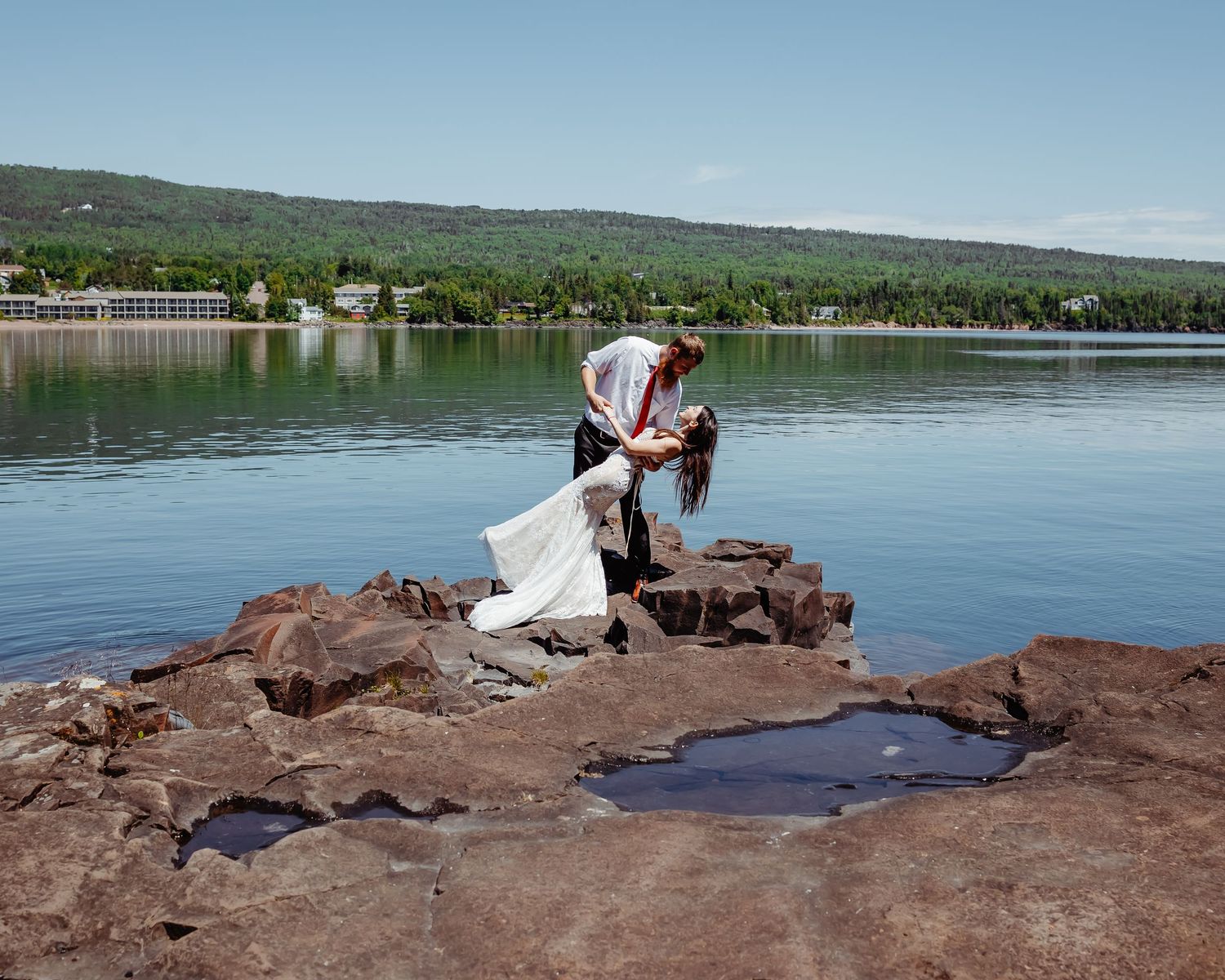 Wedding couple posing on red rock formation by serene lakeside.