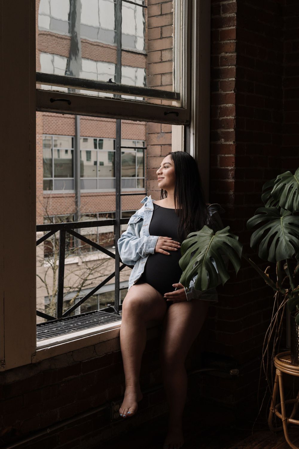 Expectant woman in casual wear sits peacefully by industrial window with houseplant and brick wall backdrop.