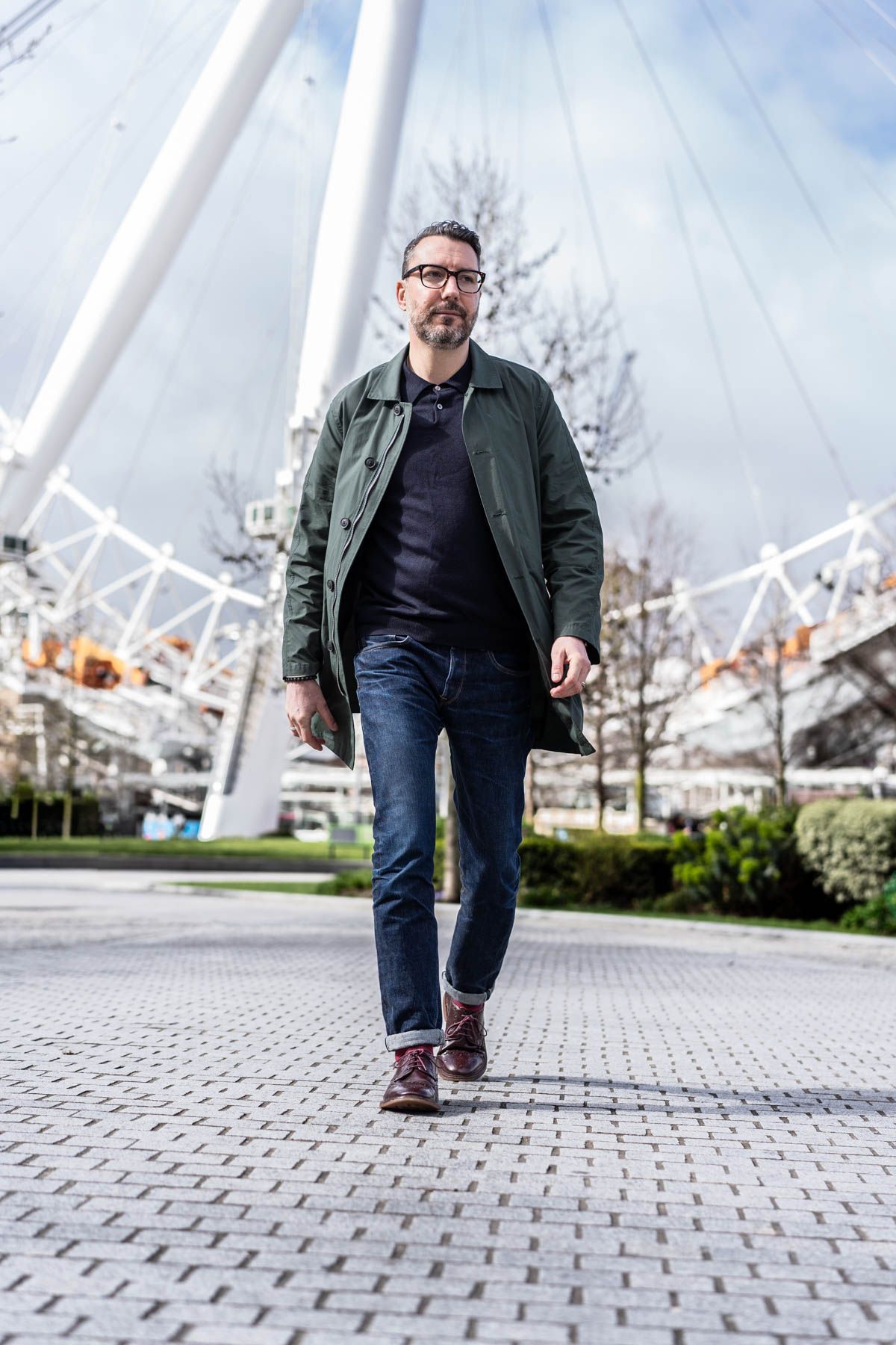 Man in casual attire with green jacket and jeans walking on paved path with London Eye in background.