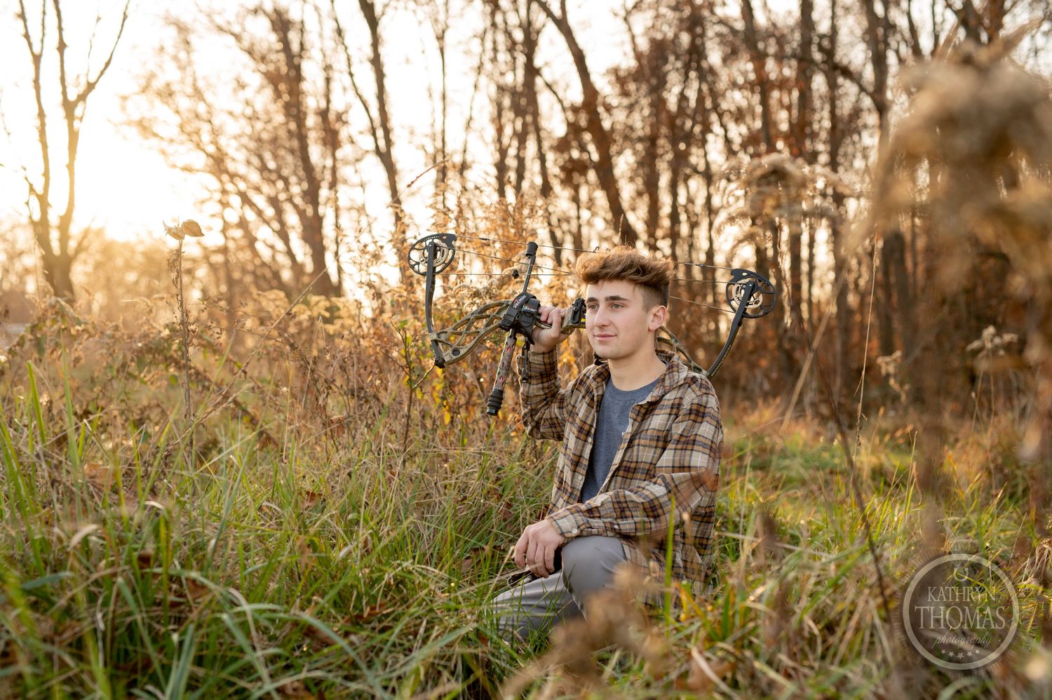 Hunter kneeling in tall grass with compound bow during autumn sunset.