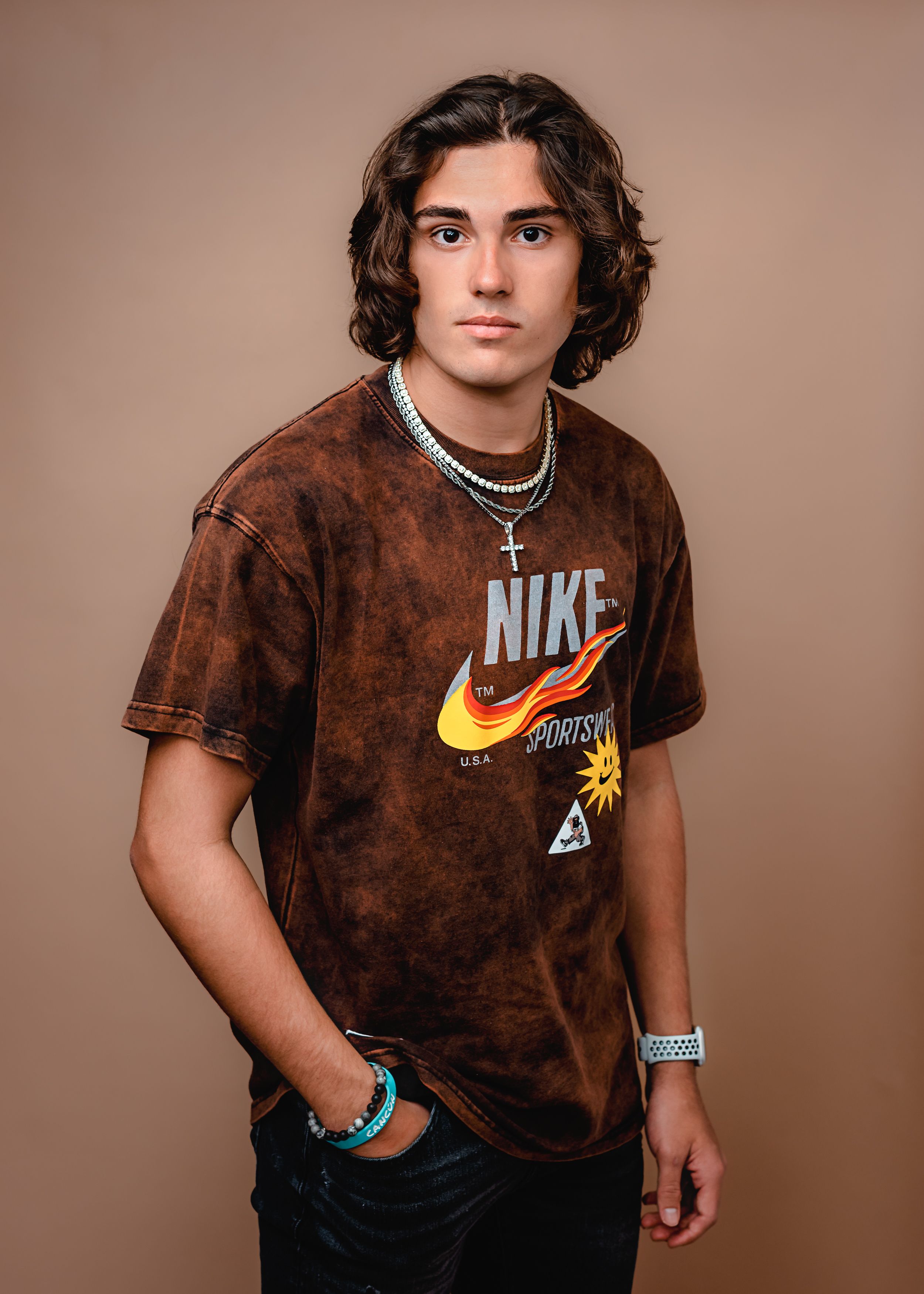 Portrait of young person wearing brown tie-dye Nike t-shirt with jewelry against neutral background.