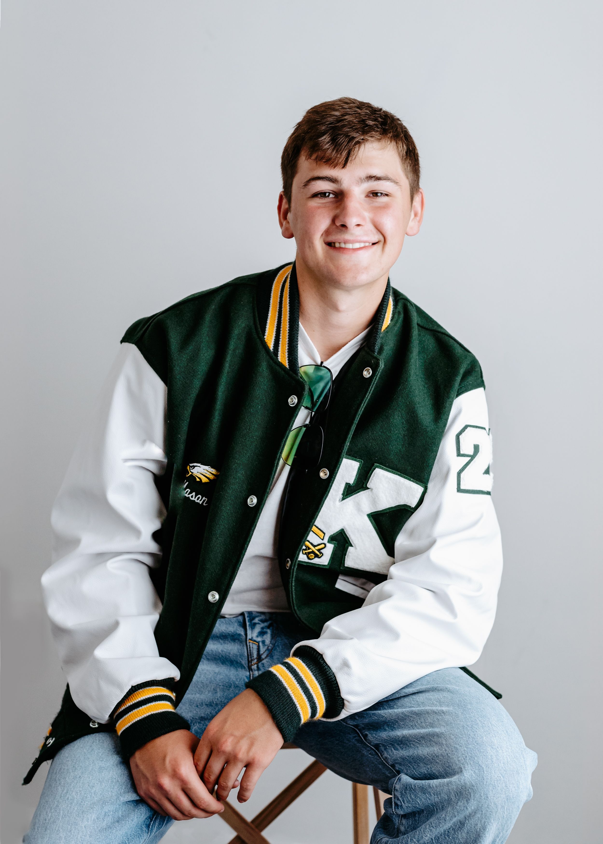 Student in a green and white varsity letterman jacket and jeans poses for a casual portrait against a light background.