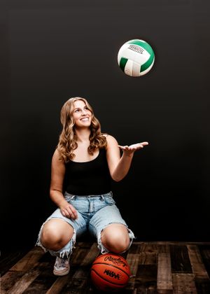 Athlete in ripped jeans plays with volleyball and basketball on wooden floor against dark background.
