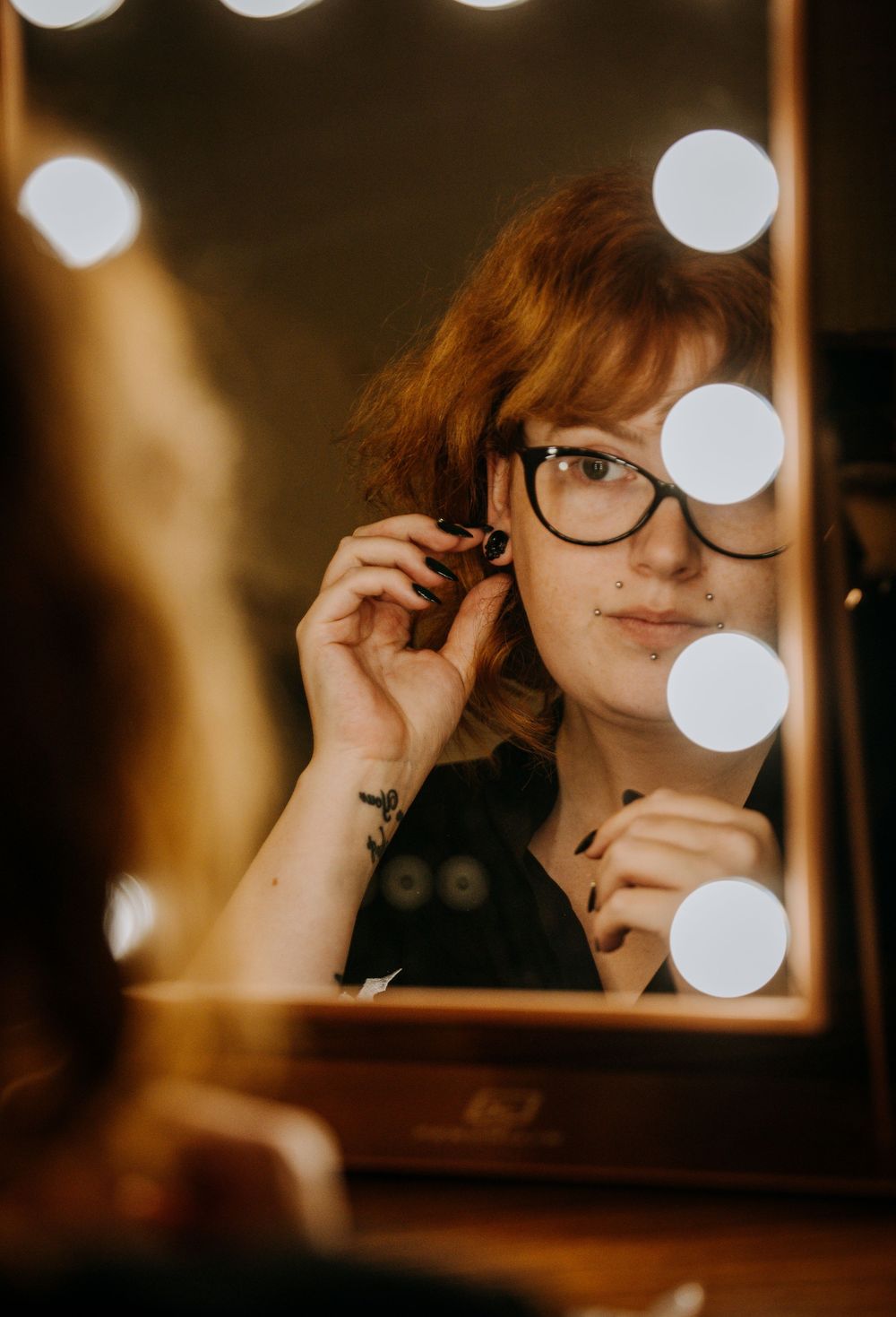 Reflection in mirror with bokeh lights shows someone wearing black-rimmed glasses in warm lighting.
