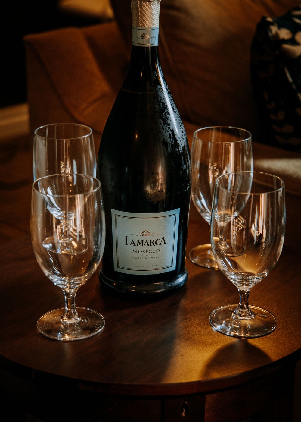 Wine glasses and a bottle of Lamarca prosecco are arranged on a dark wooden table in warm lighting.