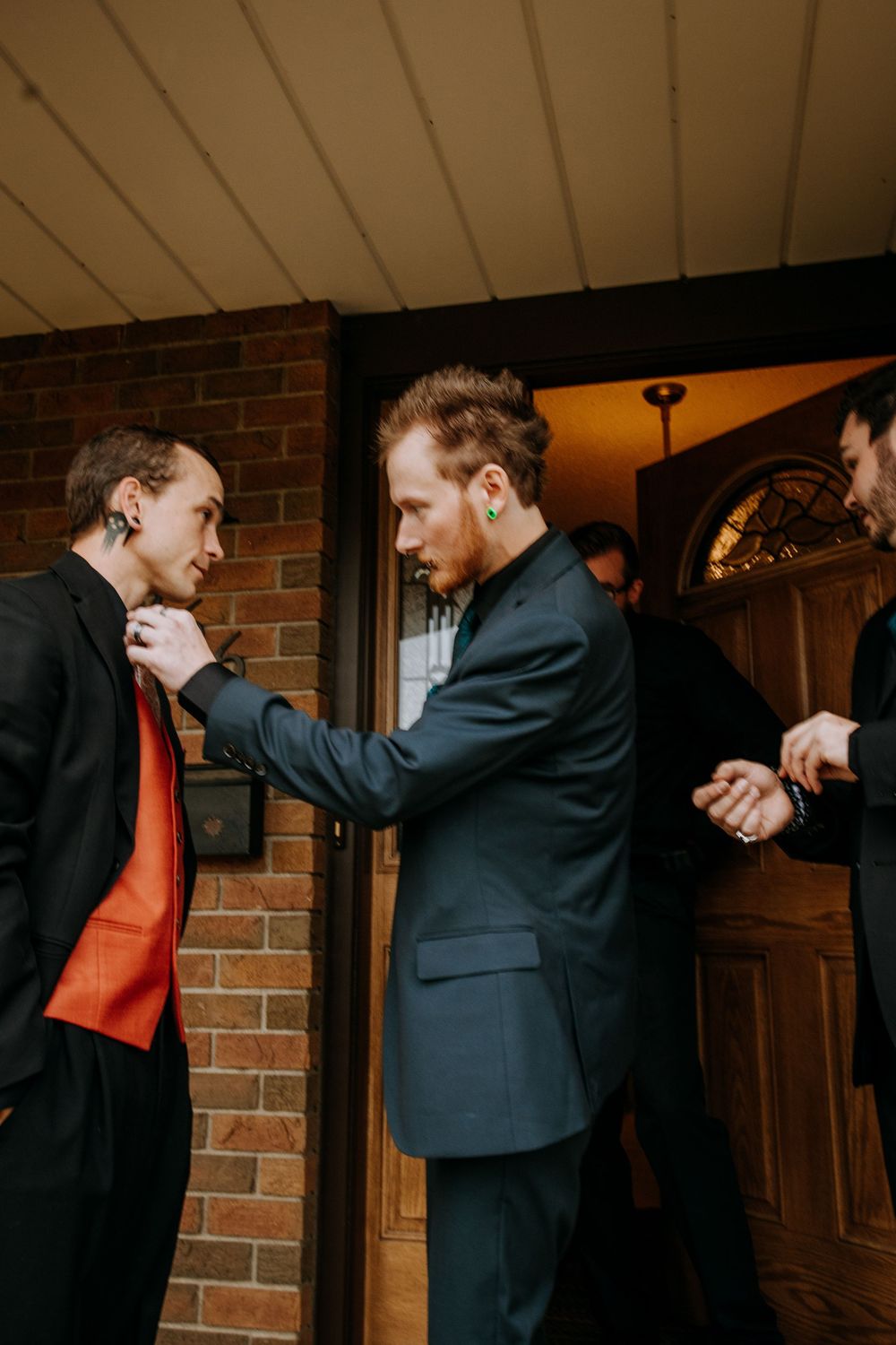 Two people in formal attire playfully interact in front of a brick wall and wooden door.