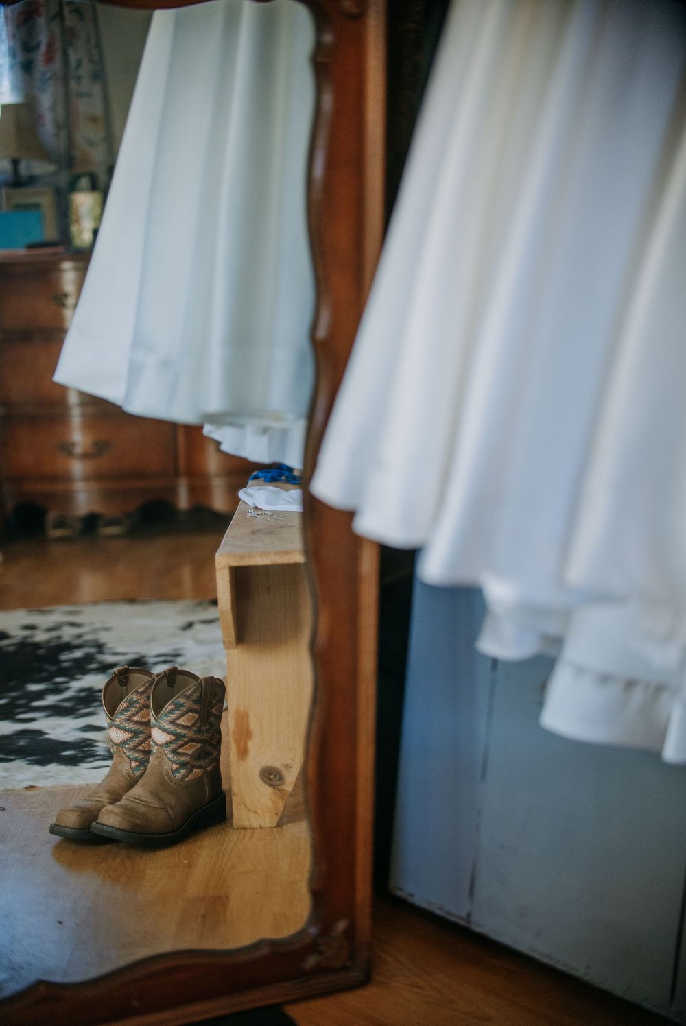 A pair of brown leather boots sits on a wooden shelf with white garments hanging in the background.